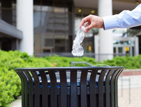 A person's hand, wearing a light blue shirt, is seen holding a crumpled clear plastic water bottle above a round, black plastic outdoor rubbish bin with vertical slats. The bin is placed on a paved surface, possibly a sidewalk or courtyard, with a background featuring modern architecture with glass windows and white structural elements. Adjacent to the bin, there are green shrubs or bushes, indicating an outdoor urban setting in the vicinity of Deptford. The scene appears to be during daylight, with natural lighting illuminating the area, emphasizing the details of the plastic bottle and the textured surface of the waste container. This image relates to waste management services, such as rubbish collection or recycling efforts, often provided by companies like Commercial Waste Removal Deptford to support local commercial and residential areas in and around the town.
