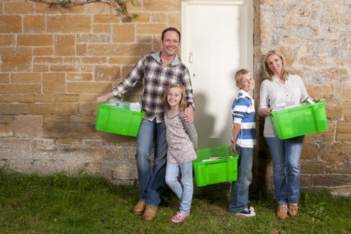 A family of four standing outside a brick building, each holding a bright green plastic storage box. The father, dressed in a checked shirt and jeans, is positioned on the left, smiling and holding a box in his right hand, with his daughter beside him also holding a box. The daughter has curly hair, wears casual clothing, and stands close to her father. To the right, a young boy in a blue and white striped shirt and jeans holds a box, looking towards the camera. Further right, the mother, wearing a white top and jeans, stands next to a textured brick wall, holding another green storage box in her left hand. The background comprises a weathered brick wall with a white door behind the family, and a patch of grass is visible at their feet. The scene suggests a casual outdoor activity related to rubbish collection or disposal, consistent with services offered by Commercial Waste Removal Deptford, serving the local postcode area.