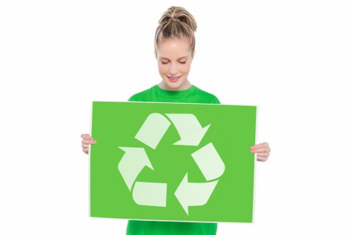 A young woman with light brown hair tied in a high ponytail, wearing a bright green long-sleeve shirt, holding a large rectangular green sign featuring a white recycling symbol composed of three chasing arrows in a triangular formation. She stands against a plain white background, smiling gently and looking down at the sign. The image emphasizes eco-friendly messages related to waste recycling and environmental responsibility, which are relevant to commercial rubbish removal services in Deptford or nearby areas.