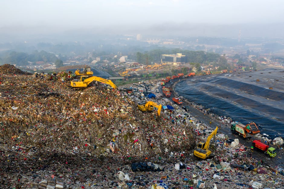 The image depicts a large landfill site filled with a mixture of waste materials including plastics, textiles, and various debris, spread unevenly across a sloped terrain. Several yellow excavators ar