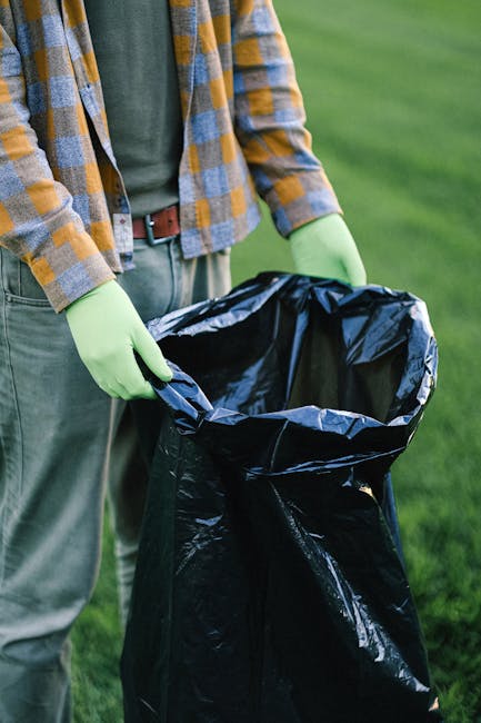 A sanitation worker wearing a yellow and red high-visibility vest is engaged in rubbish collection at the rear of a large red waste disposal vehicle, situated on a paved street. The worker is using to